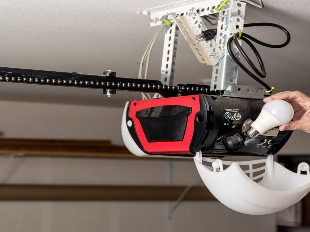 A chain-drive garage door opener mounted on the ceiling, secured with metal brackets. A person is replacing a light bulb in the opener’s built-in light fixture, which has a white plastic cover hanging open. The black power cord is looped and secured with zip ties, while the chain mechanism extends along the ceiling to operate the garage door.