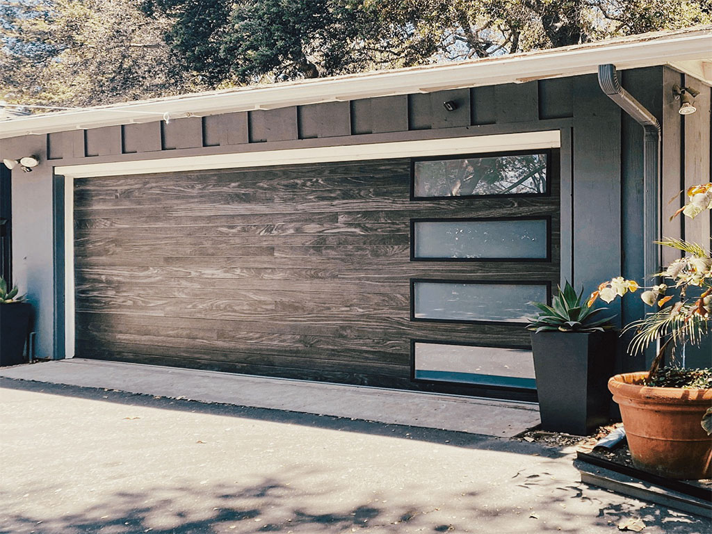 Modern wood-textured garage door with frosted glass windows on a dark-paneled house, surrounded by greenery.