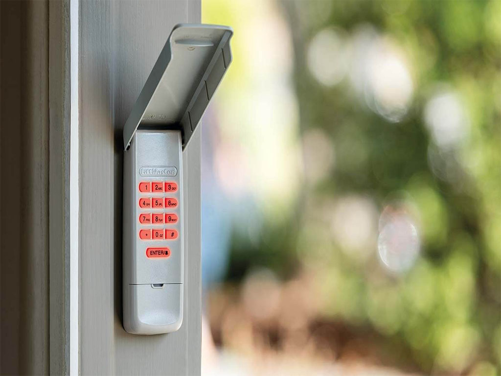 A LiftMaster wireless garage door keypad mounted on a door frame. The keypad has an open protective cover, revealing illuminated red numbers and an 'Enter' button. The background is blurred with green foliage, creating a natural outdoor setting.