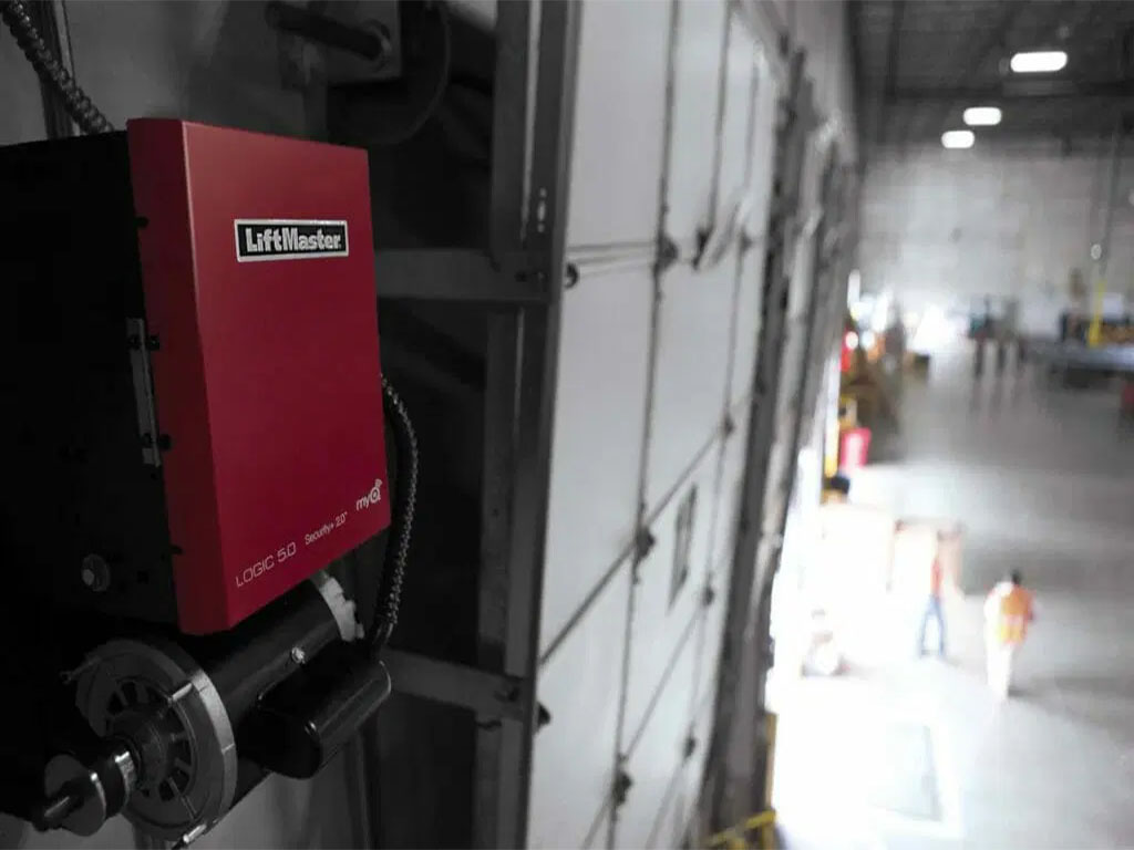 A LiftMaster commercial garage door operator installed on a sectional overhead door in a large warehouse. The red motorized unit is mounted on the side, with electrical wiring and a chain mechanism visible. The background shows an industrial setting with workers and equipment.