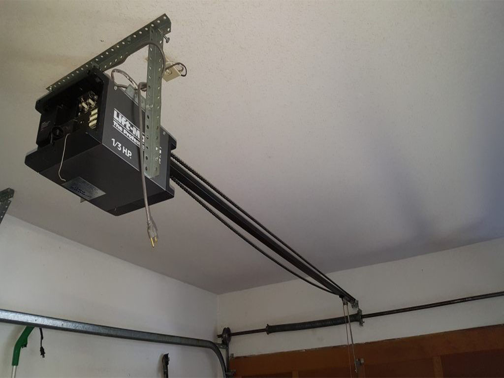 LiftMaster 1/3 HP chain-drive garage door opener mounted on the ceiling. The opener is attached to a metal bracket and rail system, with a power cord hanging down. The chain-drive mechanism extends along the ceiling, connecting to the garage door for automated operation.