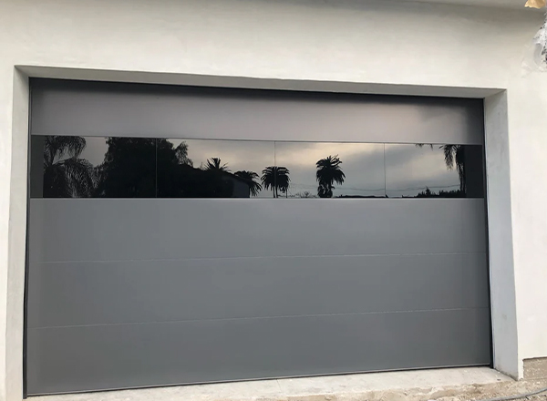 A modern gray garage door with a horizontal band of dark glass windows reflecting trees and a cloudy sky is shown. The door is installed in a light-colored stucco wall.