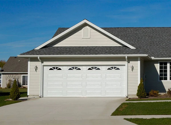 A white double garage door with arched windows is set into a beige house with a gray roof.
