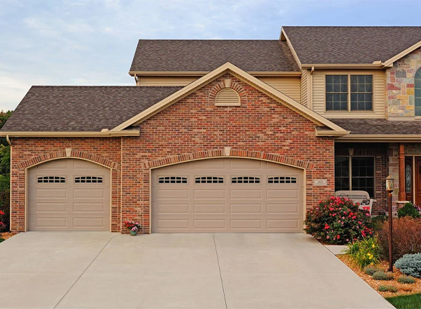 A house with a three-car attached garage is shown. The garage doors are light beige with arched tops and rectangular windows. The house is made of red brick and has a light brown roof. A concrete driveway is in front of the garage. Landscaping is visible around the house.