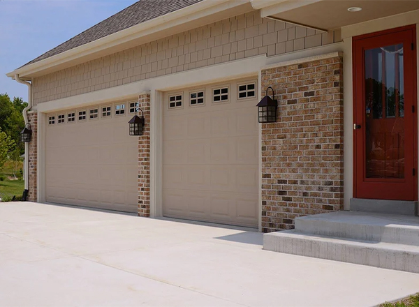 A three-car garage with beige doors and windows is shown. The garage is attached to a house with tan siding and a brick accent wall. A red door is visible on the house to the right of the garage. A light-colored concrete driveway is in front of the garage.