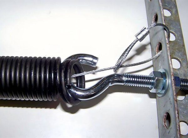 A close-up view of a garage door extension spring attached to a metal bracket. The spring is black, and the bracket is gray. A metal cable and hook are used to secure the spring to the bracket.