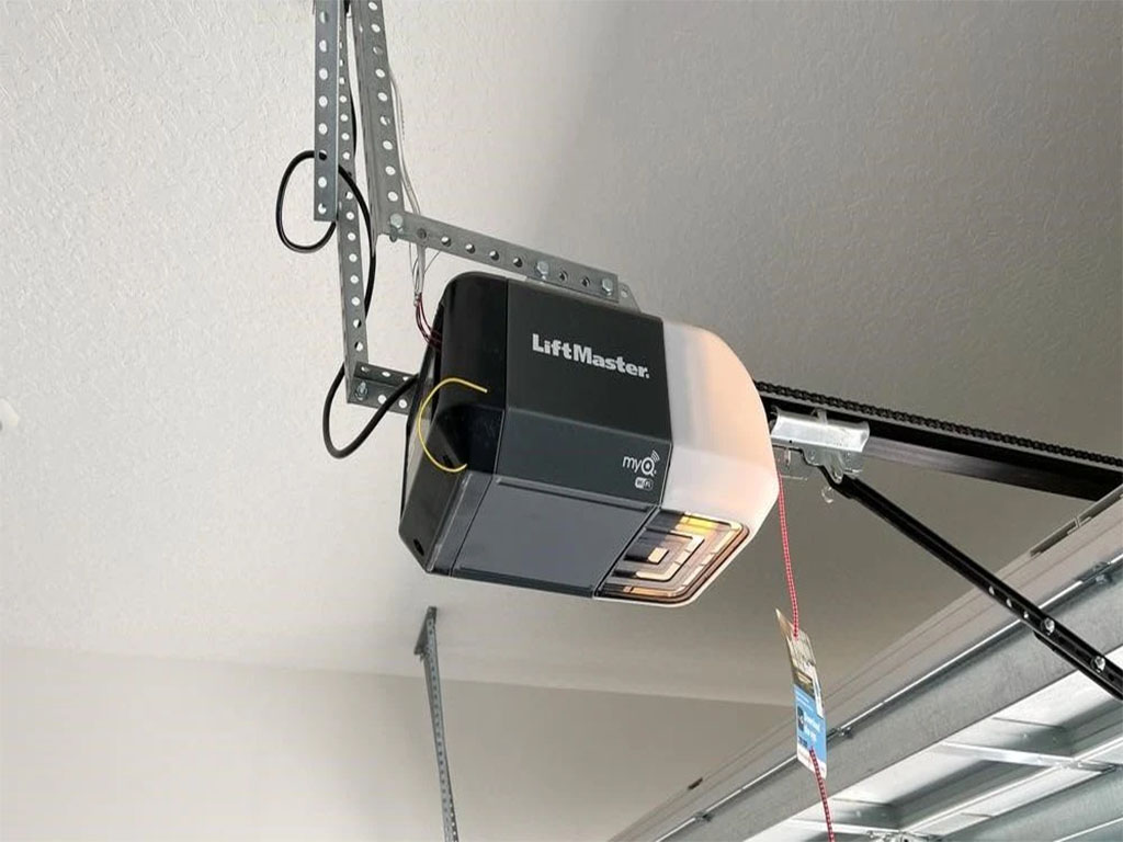 A LiftMaster Logic 5.0 commercial garage door operator installed on a sectional overhead door in a large warehouse. The red motorized unit is mounted on the side, with electrical wiring and a chain mechanism visible. The background shows an industrial setting with workers and equipment.
