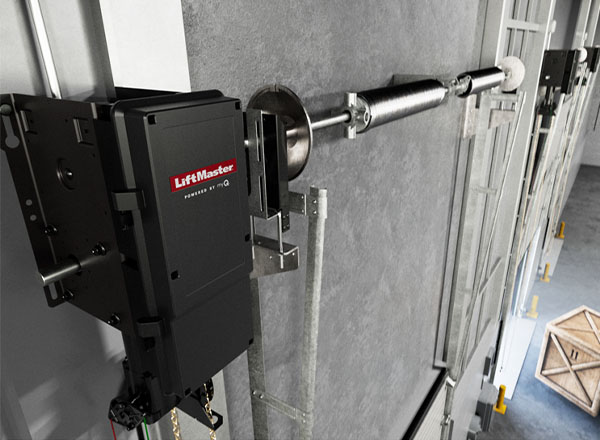 A LiftMaster commercial garage door opener is shown mounted to a concrete wall. The opener is black with a red logo. A metal roller and chain drive mechanism are connected to the opener. Part of a large industrial door is visible in the background. A wooden crate is also visible in the background.