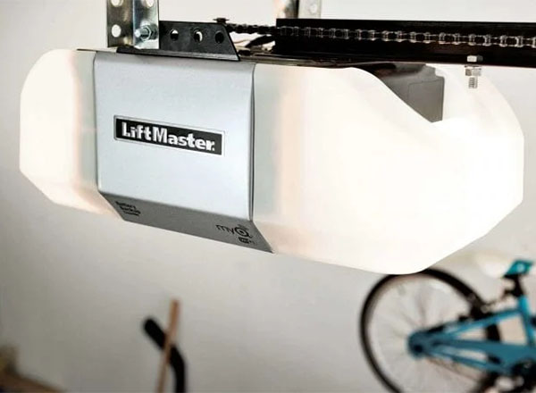 A LiftMaster garage door opener is shown mounted to a garage ceiling. The opener is primarily white with a gray top panel. A bicycle is blurred in the background.