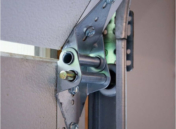 A close-up view of a garage door hinge and roller assembly. The hinge is metallic silver, and the roller is white. The hinge is attached to a light beige garage door section.
