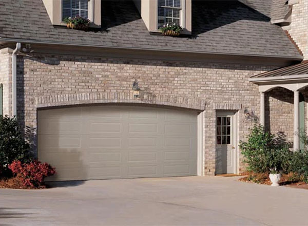 A light beige garage door with long panels is set into a brick wall.