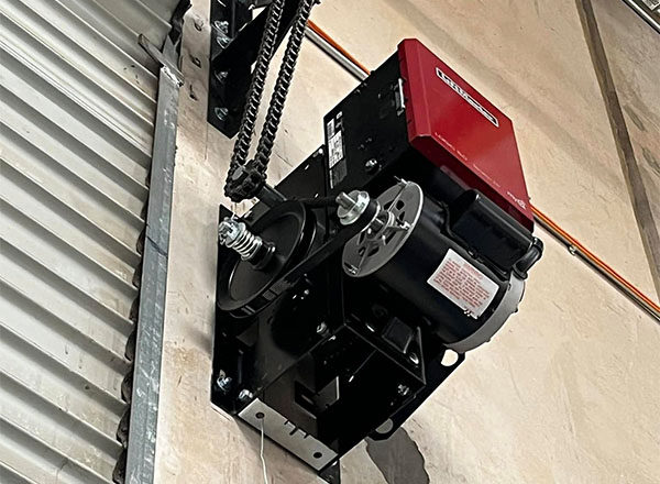 A close-up view of a commercial-style garage door opener mounted on a wall. The opener is black with a red control box. A chain drive mechanism is connected to the opener. Part of a roll-up garage door is visible in the background.