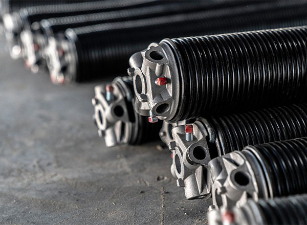 A close-up shot of several stacked garage door torsion springs. The springs are black with silver metal end caps and red accents. The springs are lying on a gray surface.